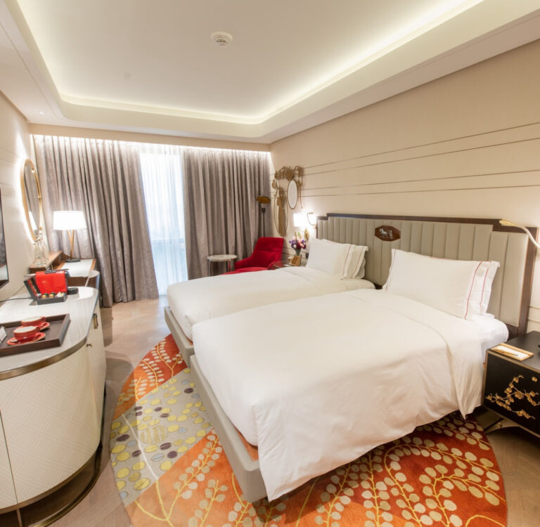 Hotel guestroom with twin beds and an orange patterned carpet at the foot of the bed